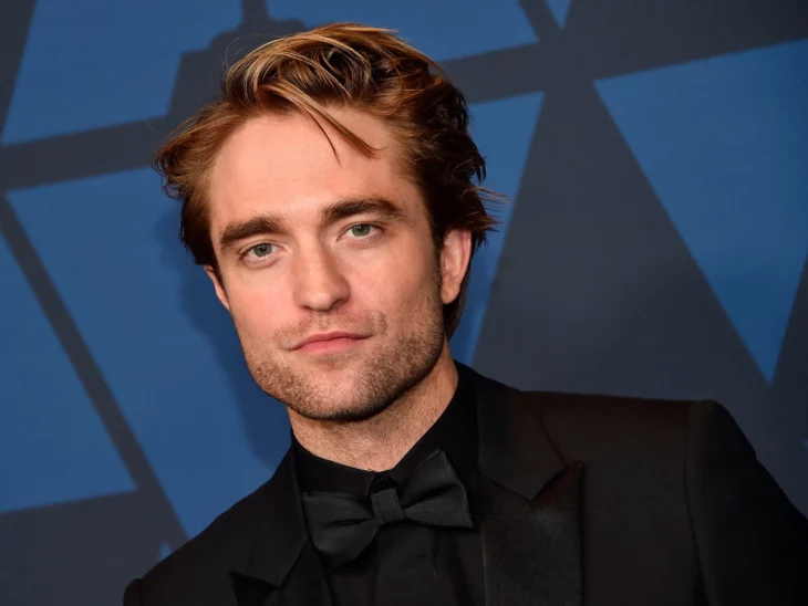 Robert Pattinson, image of his face, has short hair and a barely marked barab and mustache, wears a black jacket with a black shirt and a bow, also black