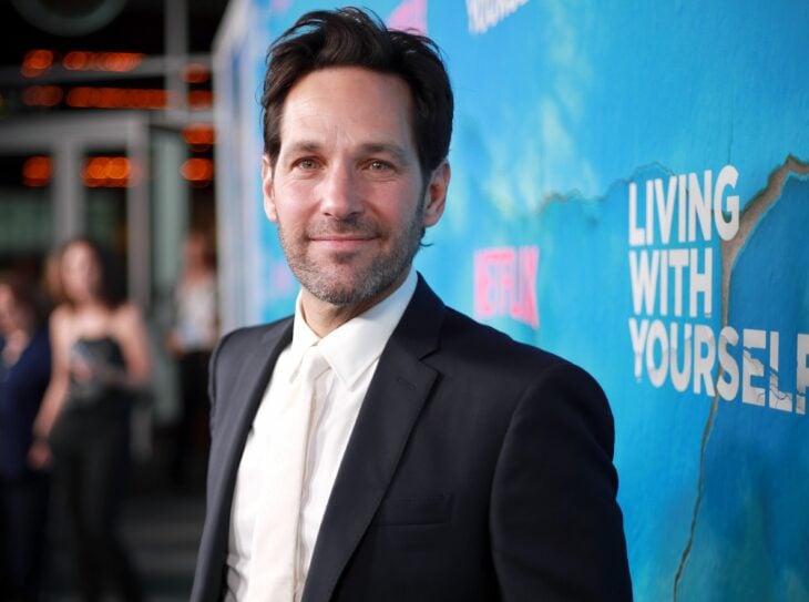 Paul Rudd smiling on a red carpet wears a black suit, a white shirt and a cream tie, the shadow of his beard and mustache can be seen 