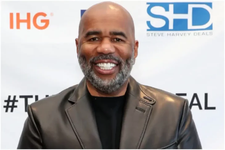 Steve Harvey smiling in the image wears a gray leather jacket and a black T-shirt his beard is long and gray 