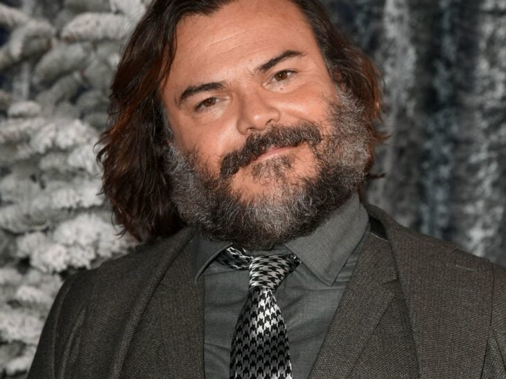 an image of Jack Black wears a gray suit with a shirt and vest, also in a gray tone, he wears a gray tie with black squares, his beard is quite long, as is his hair