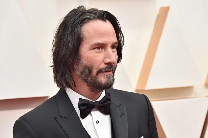 Keanu Reeves is posing on a red carpet, he is wearing a black suit with a white shirt and a black bow, his hair is a little long and his beard and mustache also look very marked