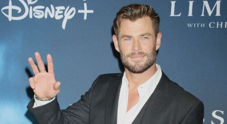 Chris Hemsworth posando en una alfombra roja lleva un saco negro y camisa blanca y saluda levantando su mano