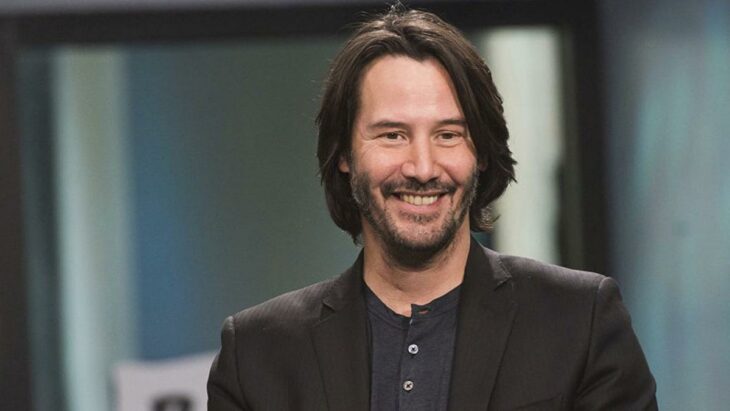 Keanu Reeves sonriendo para la cámara lleva un saco cfé y una camisa sin cuello color gris el cabello lo trae un poco largo y la barba y el bigote están crecidos