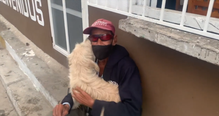 a grandpa lovingly hugs his little dog who serves as a guide and pet the little dog is cream colored and its owner wears a navy blue jacket with a red cap and sunglasses has a full beard and mustache