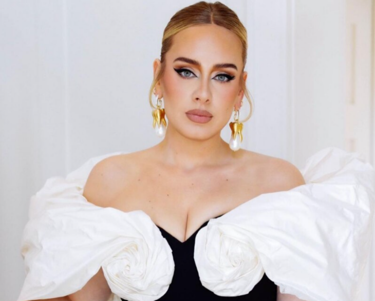 La cantante Adele posando para la cámara leva un vestido de noche con escote pronunciado en colores blanco y negro trae el cabello recogido y se aprecian unos aretes dorados en sus orejas su maquillaje está muy marcado 