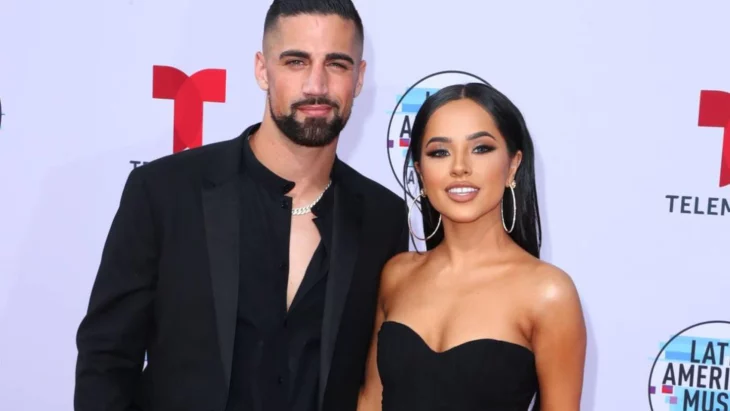 Becky G y Sebastian Lletget juntos en una alfombra roja ella lleva un vestido con escote palabra de honor en color negro el lleva un traje y camisa negros pero sin corbata ella luce dos grandes arracadas y un maquillaje pronunciado 