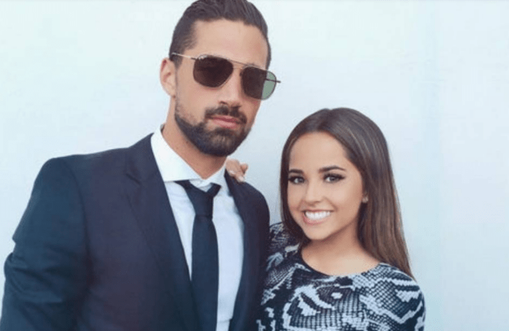 Becky G y Sebastian Lletget posando para la cámara en los primeros años de su noviazgo el lleva untraje negro con camisa blanca y corbata negra y lentes oscuros ella trae un vestido d noche plateado con negro lleva el cabello suelto y el maquillaje marcado 