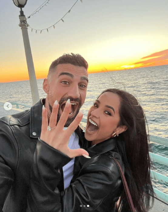 Becky G y Sebastian Lletget anunciando su compromiso con esta imagen ella luce un anillo con un enorme diamante en su dedo anular estan junto al mar vestidos con ropa casual