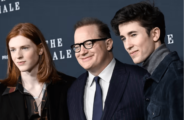 Brendan Fraser posando junto a dos de sus hijos, lleva un traje azul marino con camisa blanca y corbata también azul, los chicos visten de forma casual uno de ellos tiene el cabello pelirrojo y el otro negro