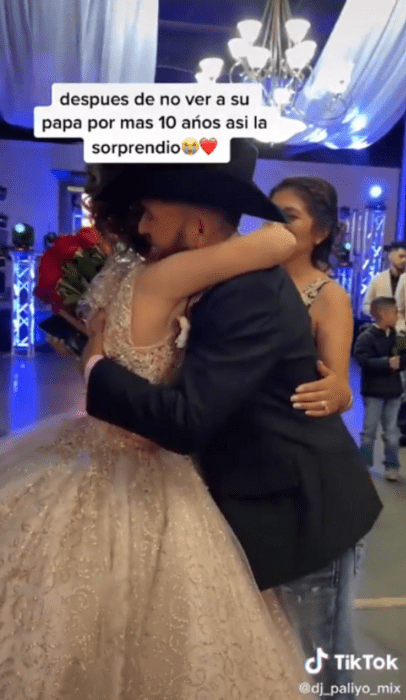 un hombre abraza a una quinceañera en una fiesta de XV años, la chica trae un ramo de rosas rojas, lleva un vestido de tul largo de color crema el hombre lleva un saco negro y una texana negra 