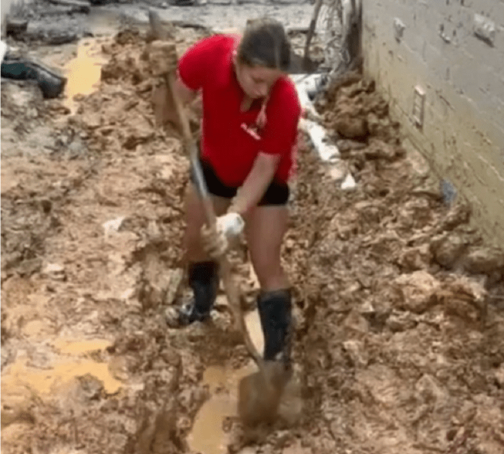 una chica realizando trabajos de plomería lleva unas botas de trabajo negras short negro y playera roja esta abriendo una zanja en el piso