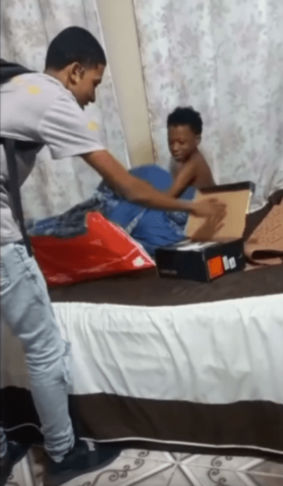 un chico entrega a su hermano unos tenis nuevos su hermano está reincorporándose de la cama mientras su hermano mayor abre una caja que contiene unos tenis nuevos el hermano mayor viste playera gris y jeans azul