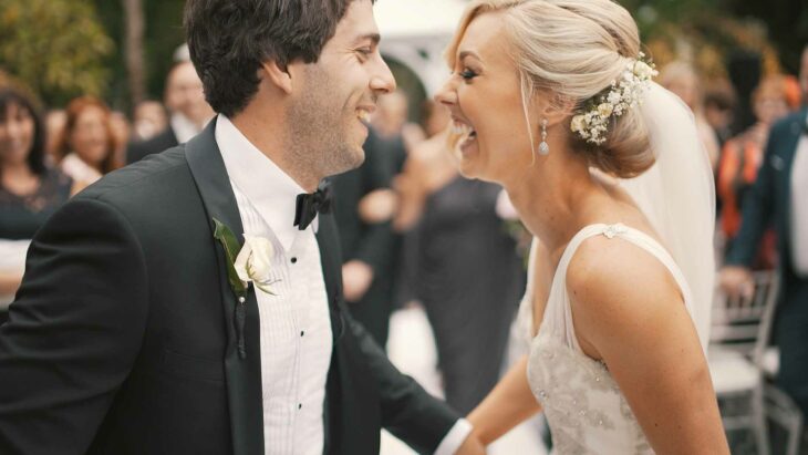 some bride and groom looking straight at each other and smiling are at their reception accompanied by their guests she wears her hair up in a bun and she has blonde hair 