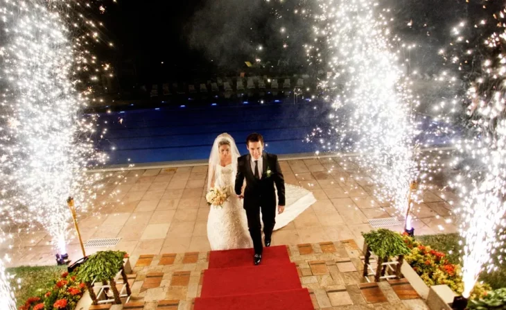 some bride and groom making their triumphant entrance to the room where their wedding reception will be held, there are cascades of fireworks around them and a red carpet