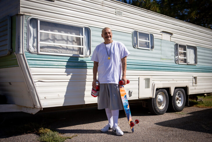 Fotografía de un hombre conocido como el "tiktoker cholo" parado frente a una casa rodante con una patineta en una mano y en la otra un jugo de arándano. 