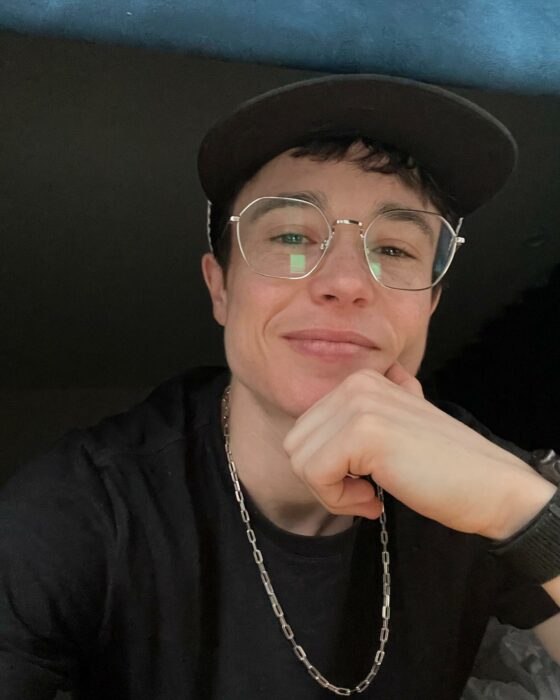 Selfie que muestra al actor canadiense Elliot Page usando gorra, lentes y una cadena sonriendo y con la mano sobre la barbilla 