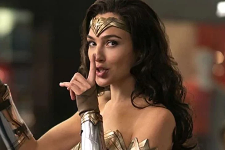 Gal Gadot personificada como la mujer maravilla lleva el cabello largo suelto y ondulado un adorno dorado cubre su frente y esta haciendo una seña con su mano como si pidiera a alguien guardar silencio