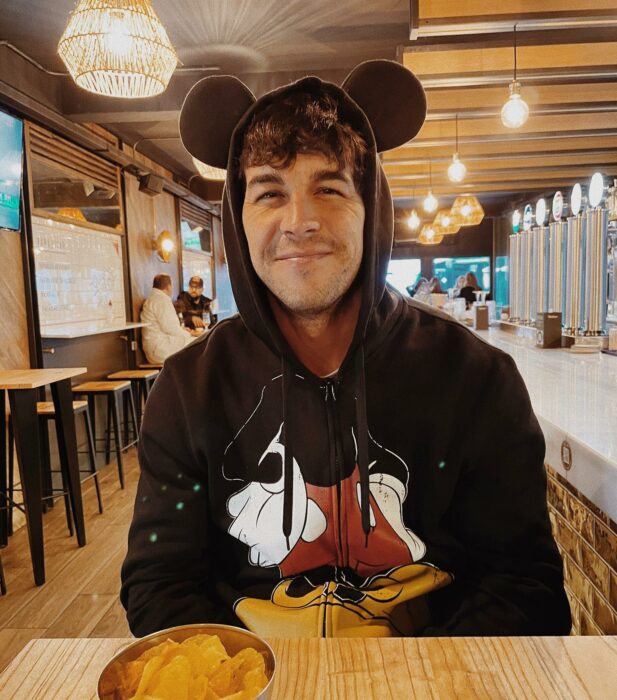 Mario Casas en la mesa de un restaurante posando con una sudadera de Mickey Mouse sonriendo 