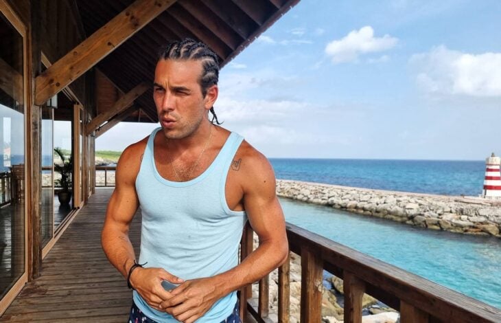 Actor Mario Casas en una casa frente a la playa con una camisa de tirantes blanca y trenzas 
