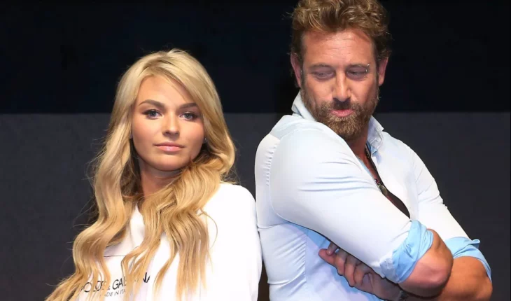 Gabriel Soto and Irina Baeva pose together and with their backs turned at a press conference for a play. She has super blonde and long hair with a white jacket 
