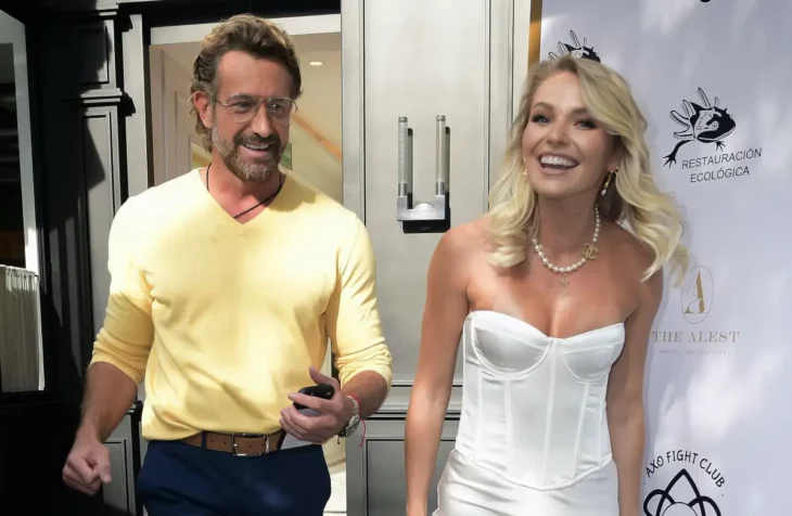 Gabriel Soto and Irina Baeva walking together, they are both smiling, she is wearing a white corset and her blond hair is loose, with a pearl necklace around her neck, he is wearing a yellow sweatshirt-type shirt and is wearing black pants