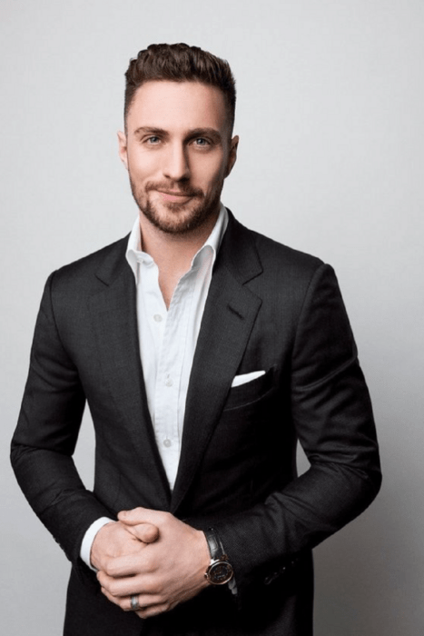 Aaron Taylor-Johnson vestido con un saco negro y camisa gris sin corbata trae el cabello corto y tiene en el rostro una mueca de sonrisa