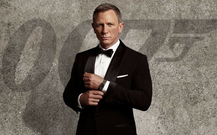 Daniel Craig personificado como Jame Bond el legendario agente 007 viste un esmoquin negro con camisa blanca lleva un moño negro en el cuello