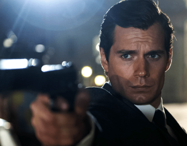 Henry Cavill en la escena de alguna película, sostiene un arma como si estuviera apuntándole a alguien lleva un traje formal negro con camisa blanca y corbata negra 