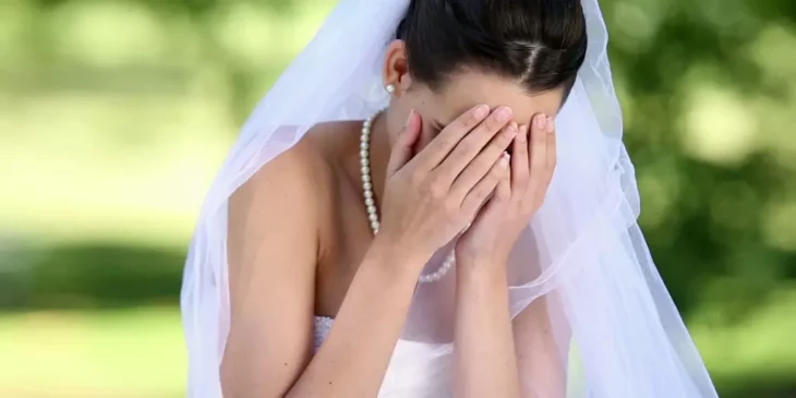 una novia vestida con un traje blanco y velo se lleva las manos al rostro como si estuviera llorando lleva el cabello recogido y un collar de perlas 