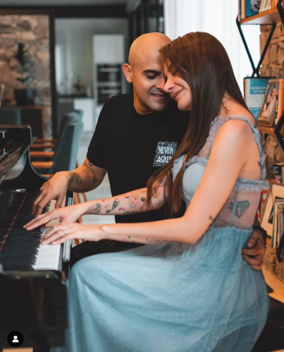 una pareja interpretando una canción al piano ella lleva un vestido vaporoso de color gris claro y tare el cabello suelto y largo él lleva una playera negra 