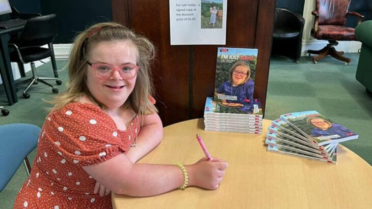 una chica con síndrome Down esta sentada junto a una mesa en una firma de libros varios ejemplares están sobre la mesa lleva un vestido naranja con flores blancas el cabello suelto y trae una pluma en la mano
