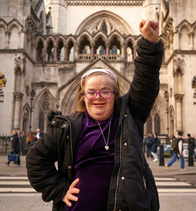 Heidi Crowter una mujer con síndrome Down esta parada en una plaza pública del Reino Unido tiene un brazo en alto como señal de lucha trae una blusa morada y una chamarra negra usa anteojos y no lleva maquillaje