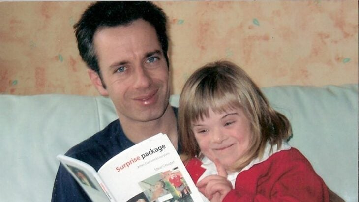 un padre leyendo un libro a su hija con síndrome Down ella está vestida de rojo y su papá lleva una playera negra ambos están sonriendo