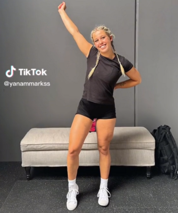 a blonde girl has one arm raised as a sign of victory, her blonde hair is tied up in two braids and she is wearing dark sportswear with shorts and a t-shirt, her tennis shoes and socks are white