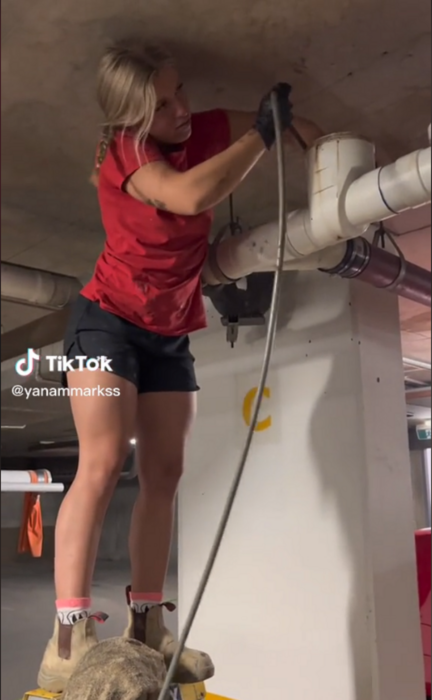 a young plumber fixing a clogged pipe has a cable in one of her hands and the other has it stuck in a pipe of the pipes she wears black shorts and a red shirt 