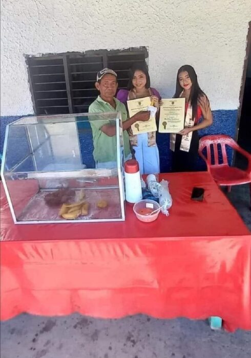 chicas acuden con un vendedor de empanadas a mostrarle sus diplomas, hay una mesa con un mantel de hule donde está colocada una pequeña vidriera sonde están las empanadas las chicas llevan sus títulos de bachillerato y los muestran en la imagen