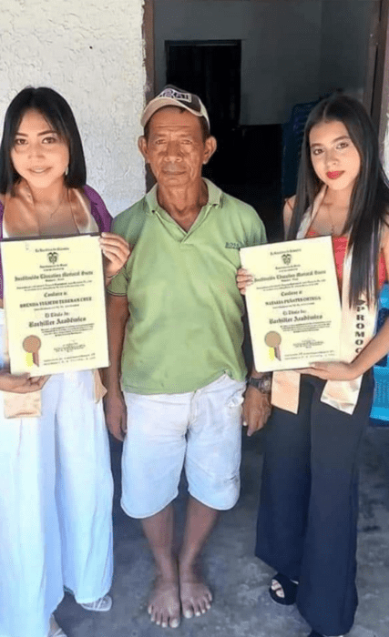 dos chicas posan junto a un humilde vendedor de empanadas llevan su título de bachillerato en las manos tienen ropa formal y el hombre que las acompaña esta humildemente vestido y no lleva zapatos 