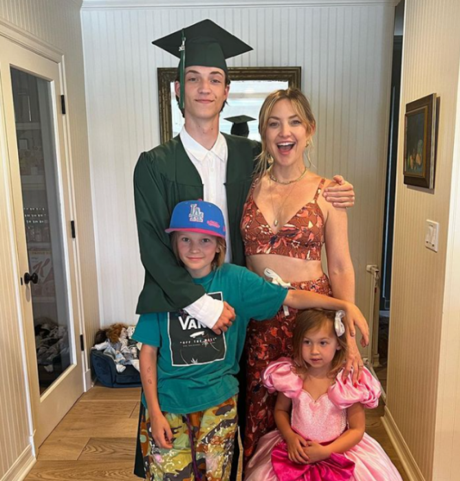 La actriz Kate Hudson con sus tres hijos, el mayor está vestido con toga y birrete pues se gradúa de la escuela el niño de 11 años trae una gorra de béisbol azul y viste deportivo y la pequeña de 4 años lleva un vestido Rosa de pincesa su madre lleva un top floreado 
