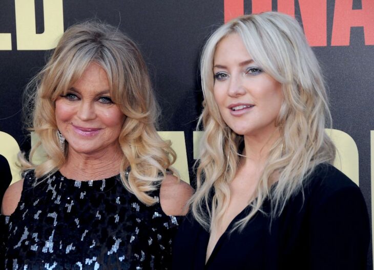 Actress Kate Hudson with her mother Goldie Hawn posing on a red carpet, both wear black suits and have loose blonde hair 