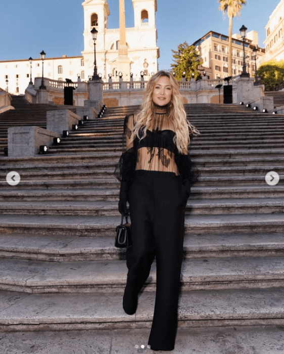 Kate Hudson posing in a black Valentino outfit wearing pants and a top under a transparent blouse and a black jacket. She is coming down some stairs and has her hair loose and long