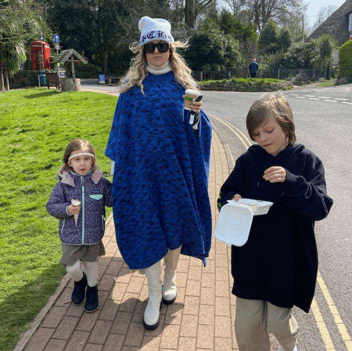 Kate Hudson with two of her children walking down the street the actress holds a coffee in her hand and is wearing a royal blue poncho with a cap and glasses her children are wearing winter clothes