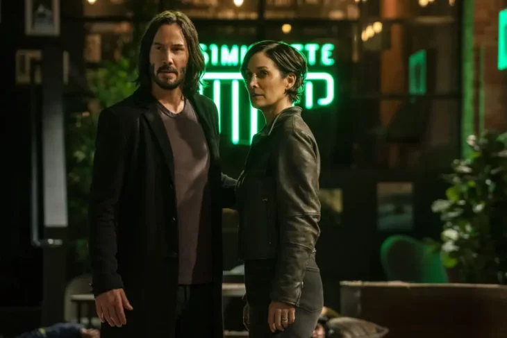 Keanu Reeves en una escena de Matrix resurrections con una de las actrices del filme lleva un saco negro y una playera gris el cabello a los hombros su acompañante lleva el cabello corto y está vestida de negro