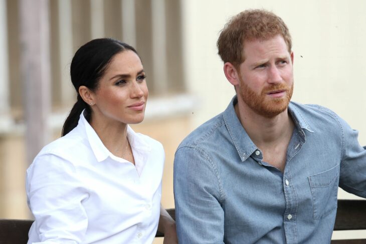 Meghan Markle and Prince Harry sitting for an interview She is wearing a white blouse with her hair in a low ponytail He is wearing a light gray shirt 