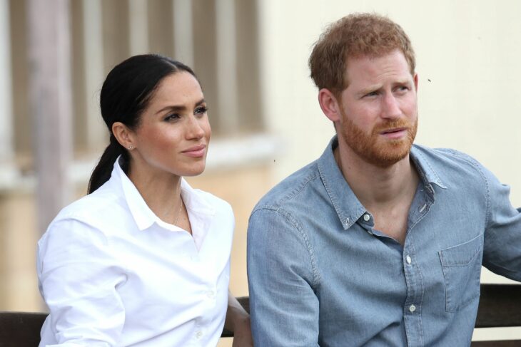 Meghan Markle y el príncipe Harry sentados dando una entrevista ella lleva una blusa blanca con el cabello recogido en una coleta baja el lleva una camisa gris claro 