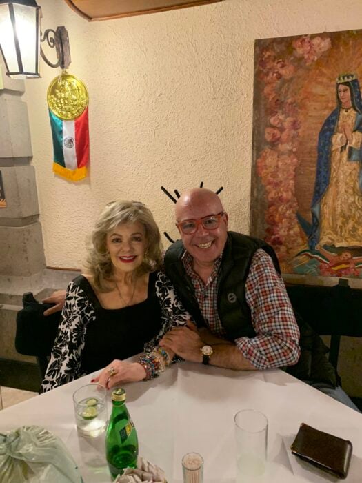 Gina Romand posando junto a su hijo Gabriel Varela en la mesa de un restaurante con un cuadro de la virgen de Guadalupe de fondo 