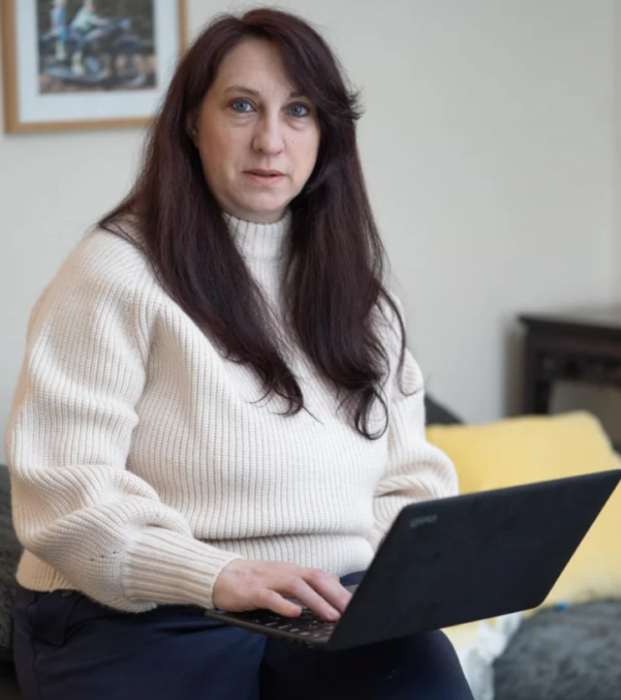 una mujer sentada sostiene en las piernas su laptop lleva un suéter crema y el cabello largo y lacio en tono castaño 