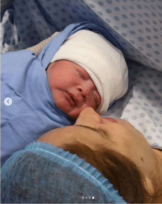 un bebesito puesto a un lado de su madre en el momento de nacer, aún lleva ropa de hospital y la cabecita cubierta con un vendaje blanco su madre trae un gorro azul 