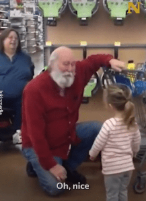 un hombre se arrodilla junto a una pequeña para charlar con ella el lleva un suéter rojo y barba larga blanca ella lleva ropa informal y una coleta están en un supermercado