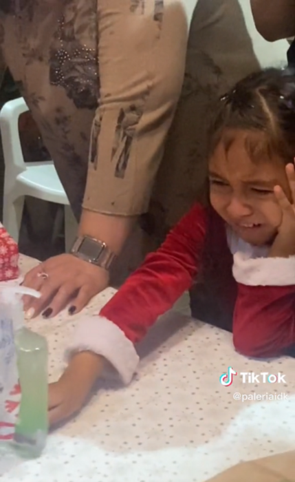 niña llora mientras sostiene con una mano un gel antibacteral que está sobre la mesa la pequeña lleva un vestido de Santa Claus