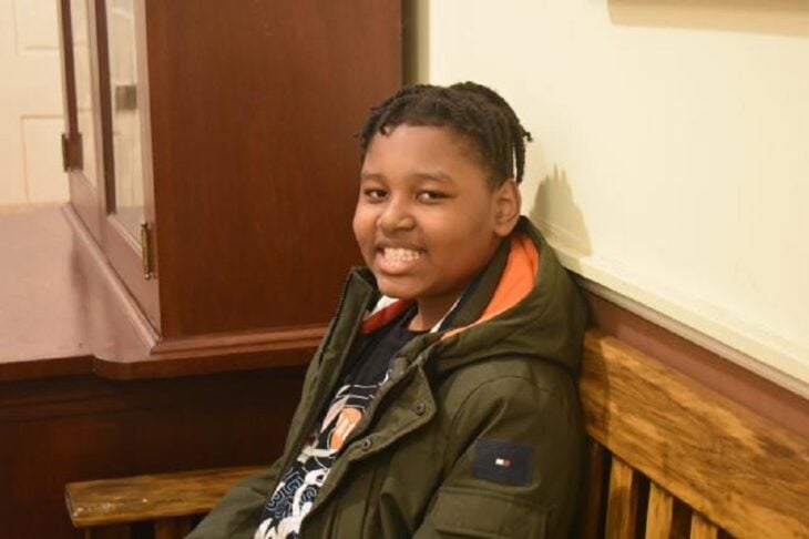 an african american boy smiles at the camera he is sitting on an office bench he is wearing a soldier green jacket and has short hair